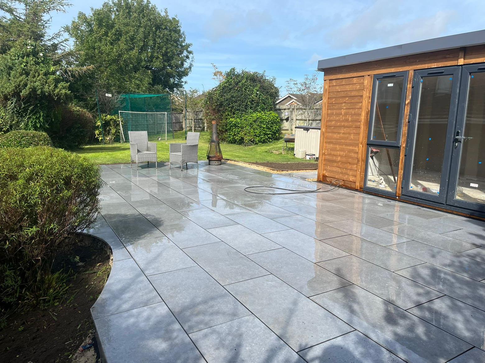A garden featuring a newly laid, shiny gray patio using expert Patio Laying, two chairs by a chiminea, a wooden shed with glass doors, green lawn, trees, and a netted play area in the background.