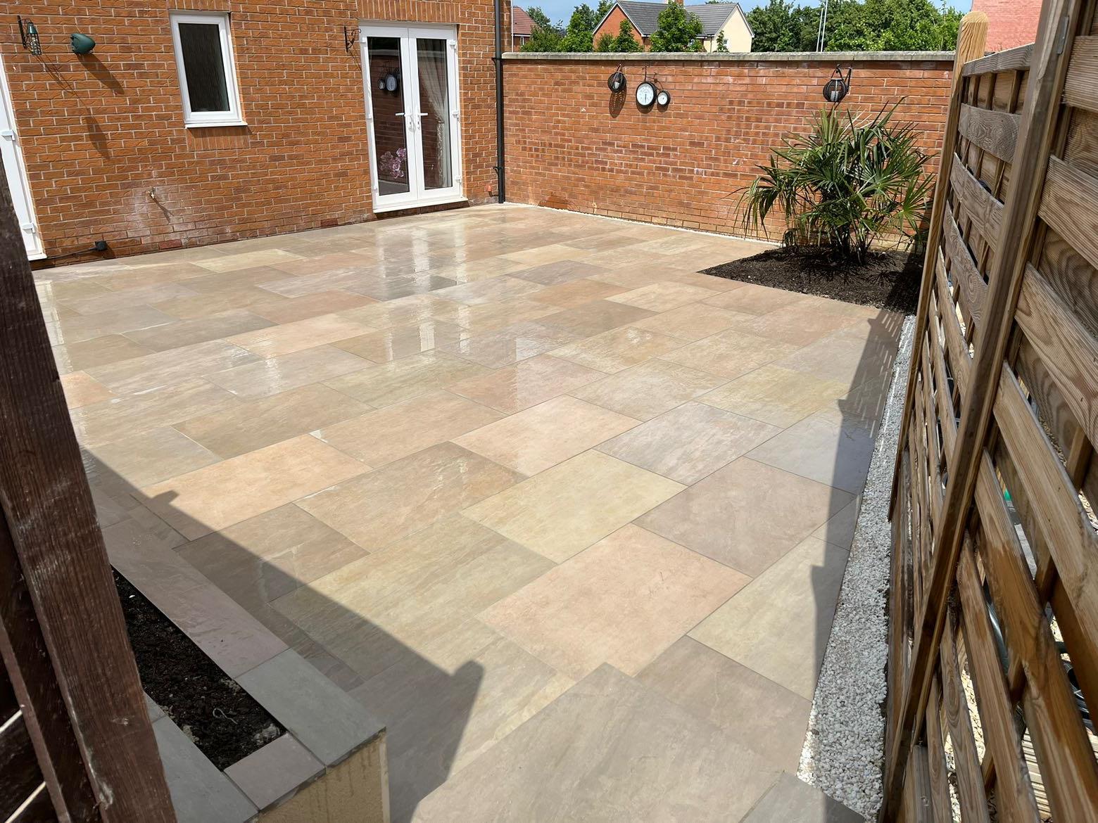 A spacious backyard patio with large, light-colored stone tiles, a small landscaped area with green plants, brick walls, block paving accents, a wooden fence, and a glass door leading into the house.