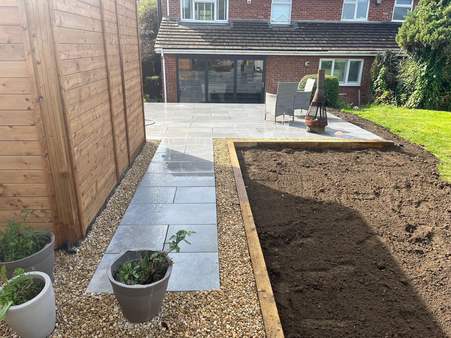 A backyard featuring expert patio laying with a paved walkway, wooden fence on the left, raised soil bed on the right, and a stylish patio area with chairs and fire pit in front of a modern house with large glass doors.