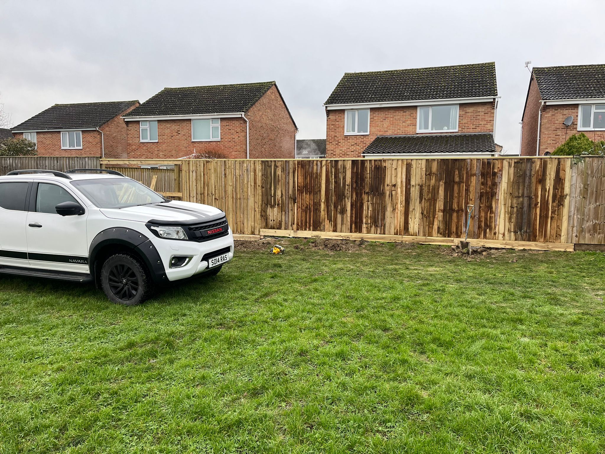 A white pickup truck is parked on grass in front of a wooden fence, with two brick houses and cloudy sky in the background—ideal for projects like patio laying or tarmac driveways.