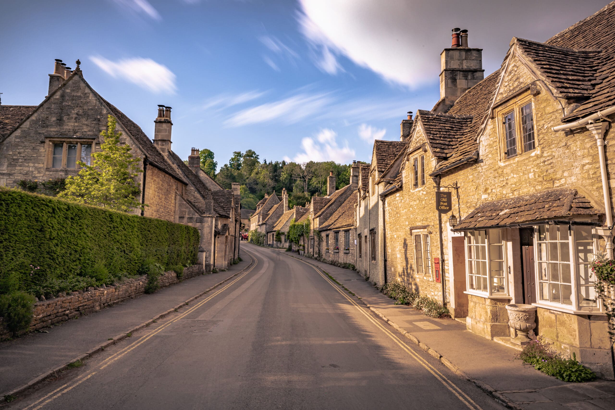 A quiet, curved street with charming stone cottages and lush greenery features expertly crafted tarmac driveways in a picturesque English village under a partly cloudy blue sky.