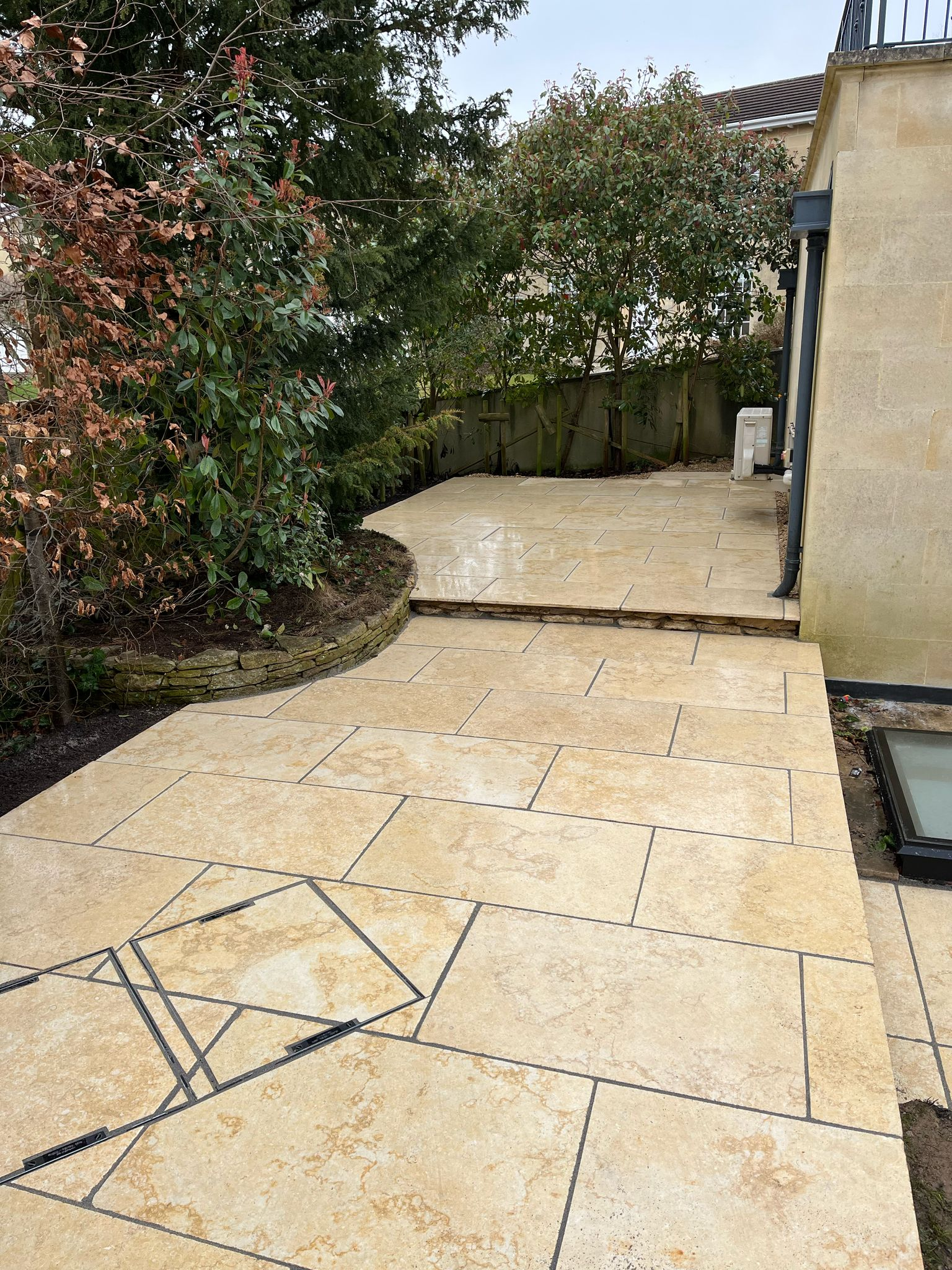 A paved stone walkway and patio, crafted with geometric tile patterns and expert patio laying, leads past a garden with trees and shrubs beside a beige stone building on a cloudy day. The tiles are light tan with thin dark lines.