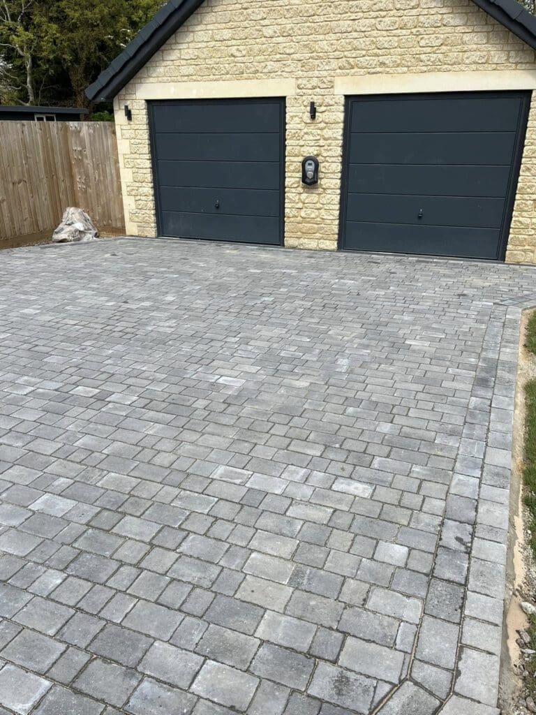A driveway made of gray block paving stones leads up to a double garage with two dark-colored doors and a stone exterior. A wooden fence and some greenery, perfect for any landscaper, are visible on the left side.