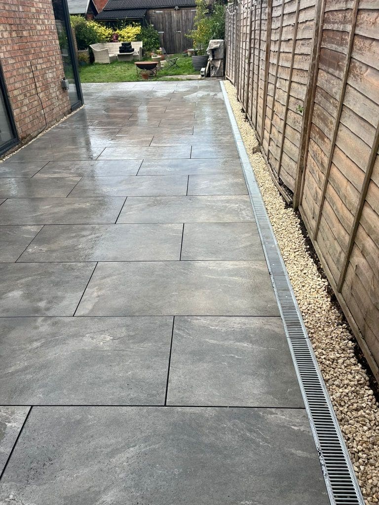 A clean, wet stone patio with large grey tiles runs alongside a wooden fence and a house. A drainage grate lines the edge, and a strip of small white stones borders the fence. Grass and garden items are visible at the far end.