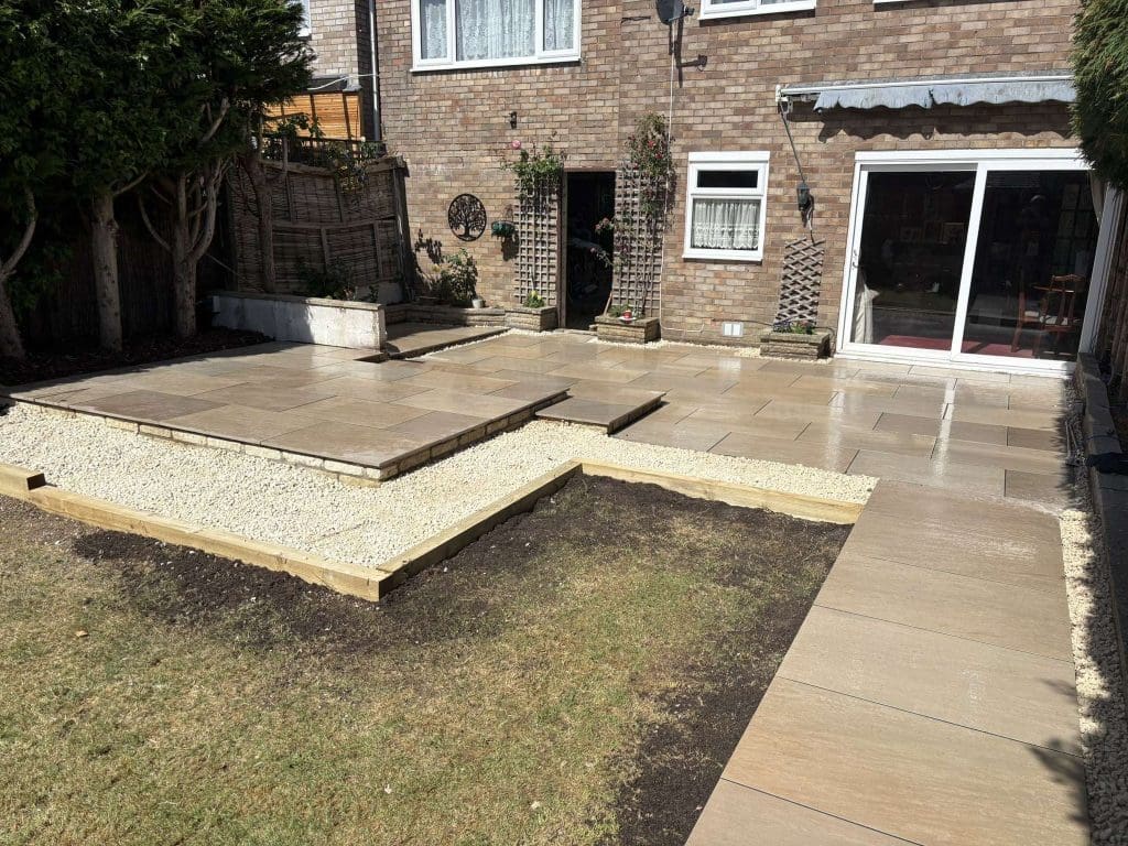 A backyard with newly installed stone patio steps, light gravel borders, wooden edging, and patches of bare soil and grass, next to a brick house with large glass sliding doors and windows.