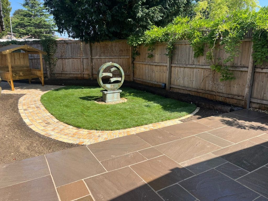 A well-kept backyard with a circular green lawn, a stone sculpture in the center, a wooden bench on the left, stone paving in the foreground, and a wooden fence with green vines along the back.