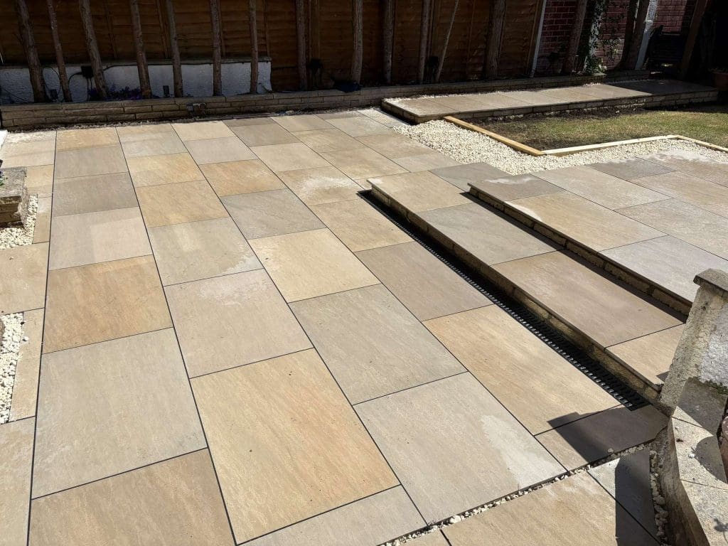 A backyard patio features large rectangular stone tiles in beige and tan tones, arranged in a staggered pattern with a narrow drainage channel running through the middle. The patio is bordered by gravel and wooden fencing.
