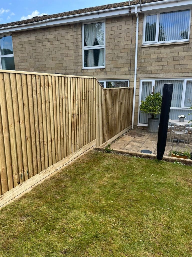 A backyard with a new wooden fence, green lawn, and a paved patio area with outdoor furniture and potted plants outside a two-story brick house.