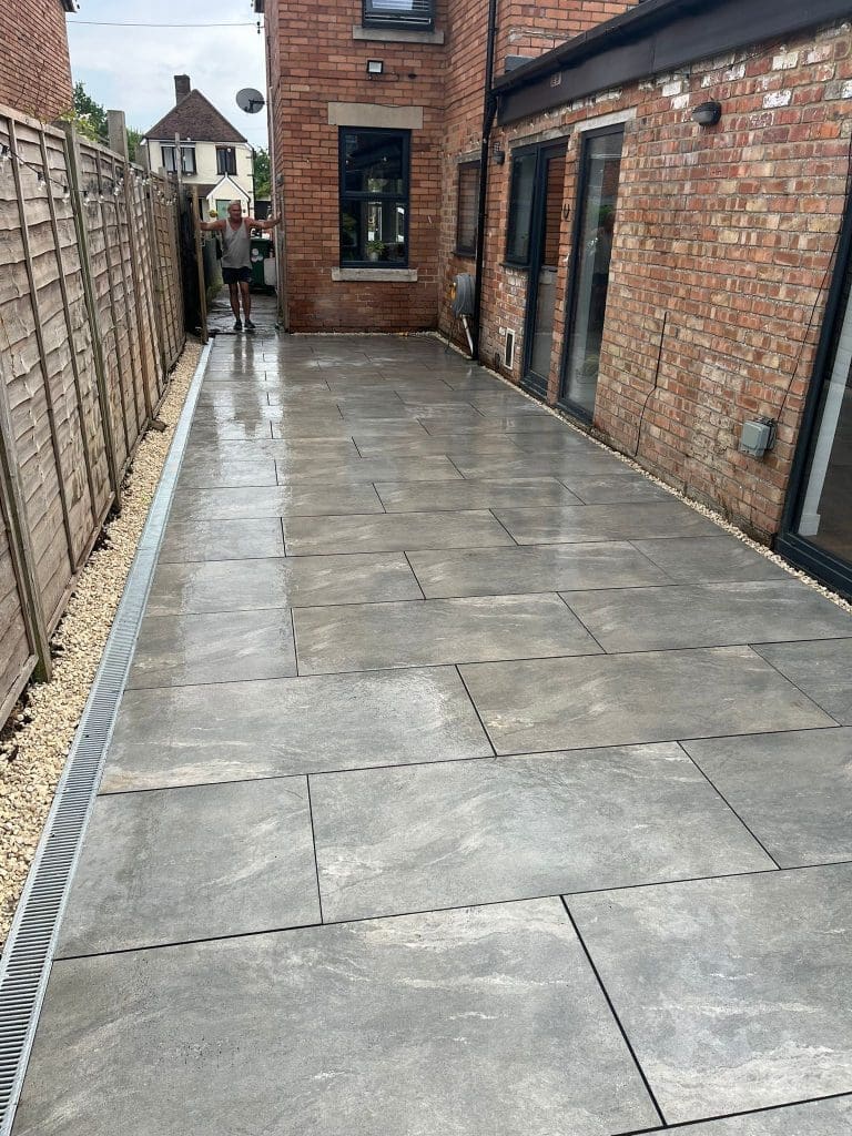 A newly paved patio with large gray tiles runs alongside a brick house. A man stands at the far end near a fence, with the patio and house reflecting some light after rain or cleaning.