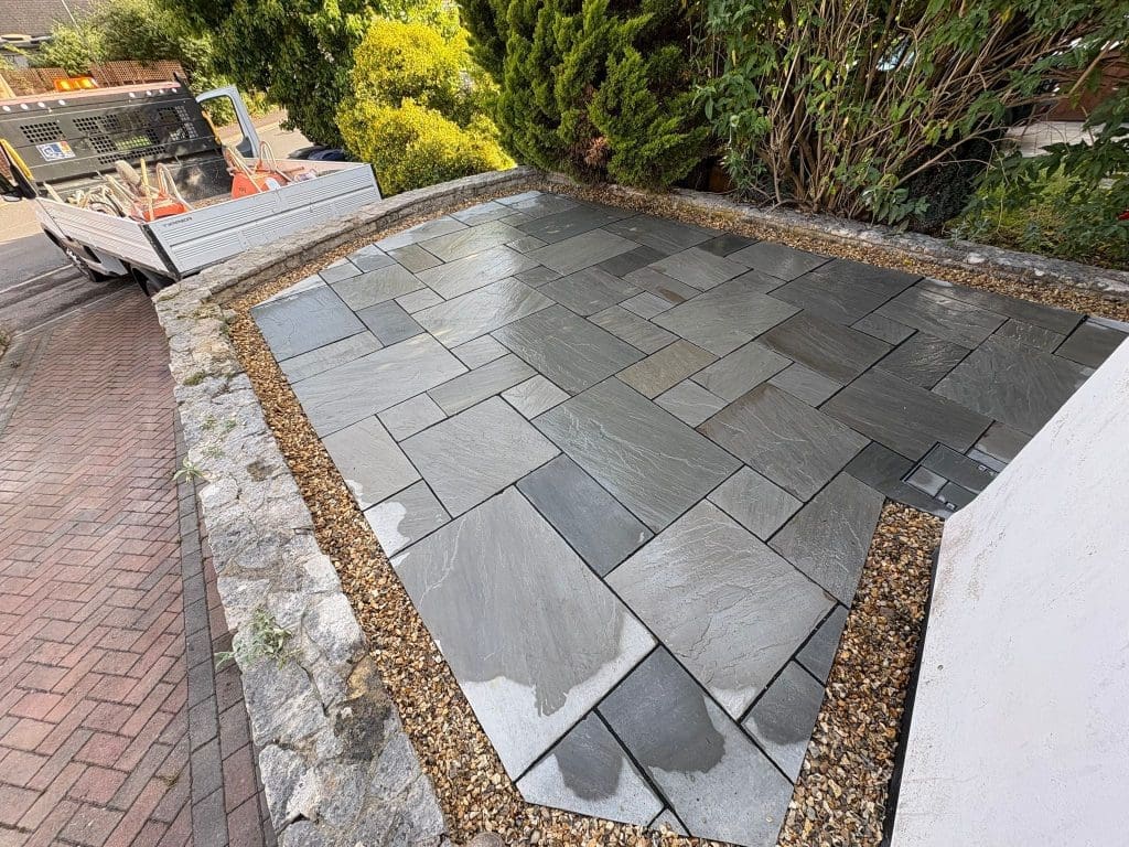 A freshly laid slate patio with rectangular and square tiles in a geometric pattern, bordered by gravel and stones. A white work truck with tools is parked on the adjacent sloped driveway. Green shrubs and trees surround the area.
