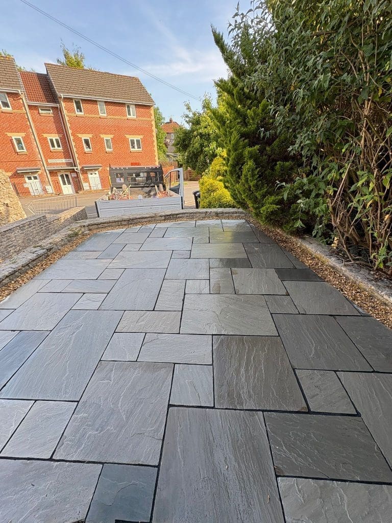 Freshly laid slate patio tiles on a backyard terrace, bordered by gravel and green bushes on one side, with red-brick houses and a clear blue sky in the background.