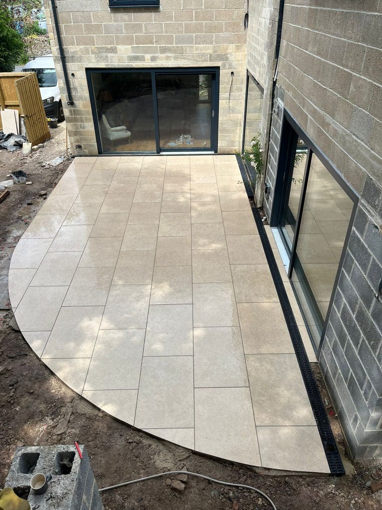 A newly installed beige tiled patio with a curved edge, situated between two stone buildings with large glass sliding doors. Construction materials and unfinished ground are visible around the patio.