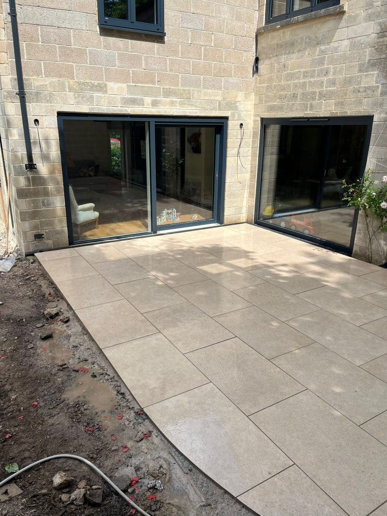 A newly installed tan tile patio outside a modern stone house with large glass sliding doors. The patio is clean and unfinished ground surrounds the area, indicating ongoing construction or landscaping.