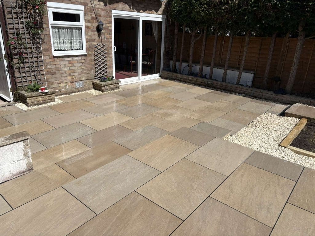A backyard patio with light brown rectangular paving stones, bordered by gravel and small garden beds, next to a brick house with sliding glass doors and a window with white lace curtains.