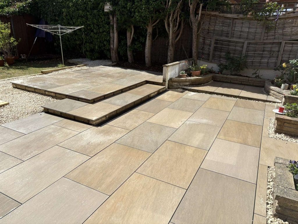 A backyard with newly laid beige stone patio tiles, raised steps, gravel borders, potted plants, a wooden fence, drying rack, and trees along the back, partially shaded by sunlight.