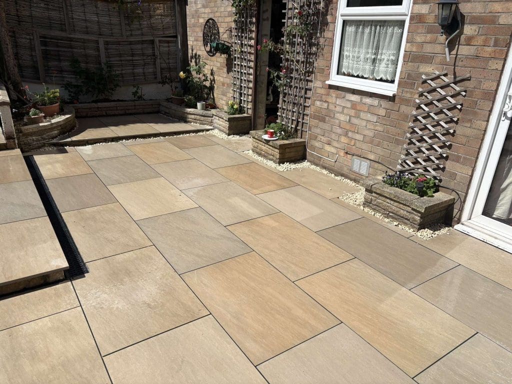 A freshly-laid patio with large beige and tan paving slabs, bordered by white gravel and raised plant beds, situated next to a brick house with windows and decorative trellises.
