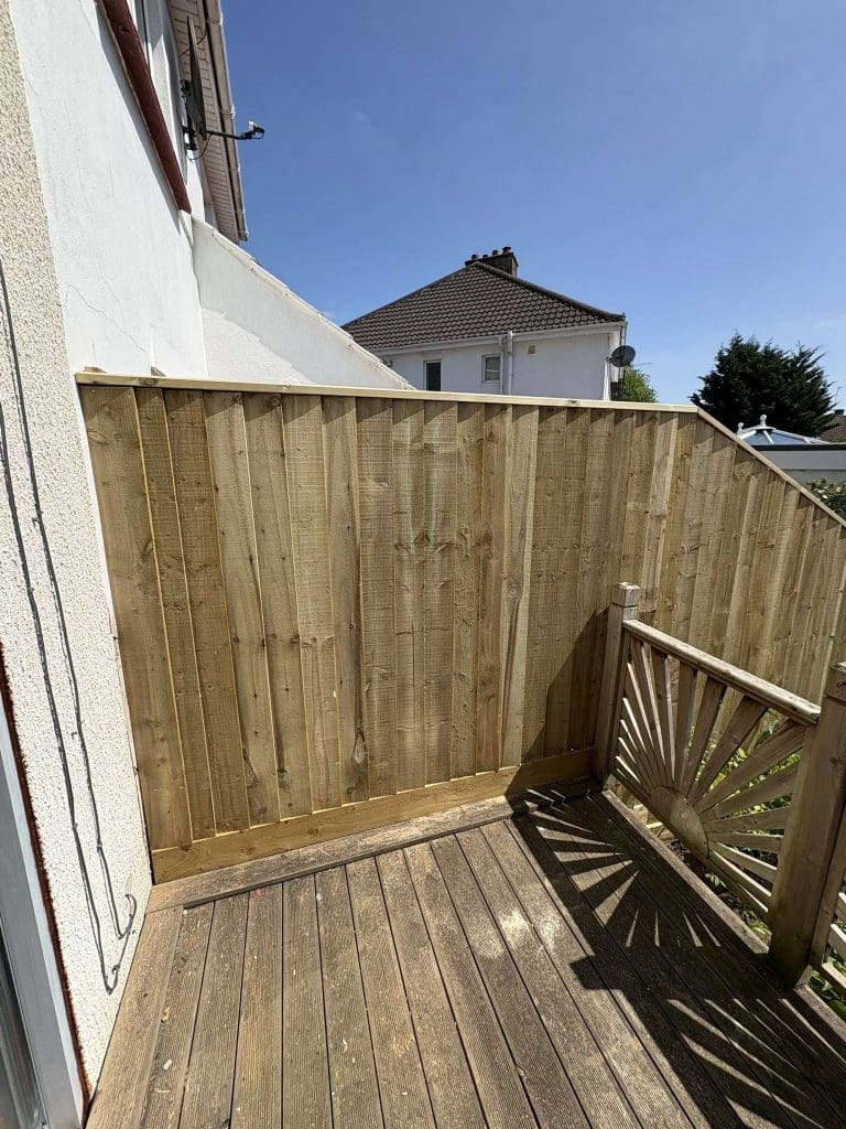 A wooden deck with a new, tall wooden privacy fence attached to the side of a house, under a clear blue sky. Adjacent is a smaller decorative wooden gate with a sunburst design.