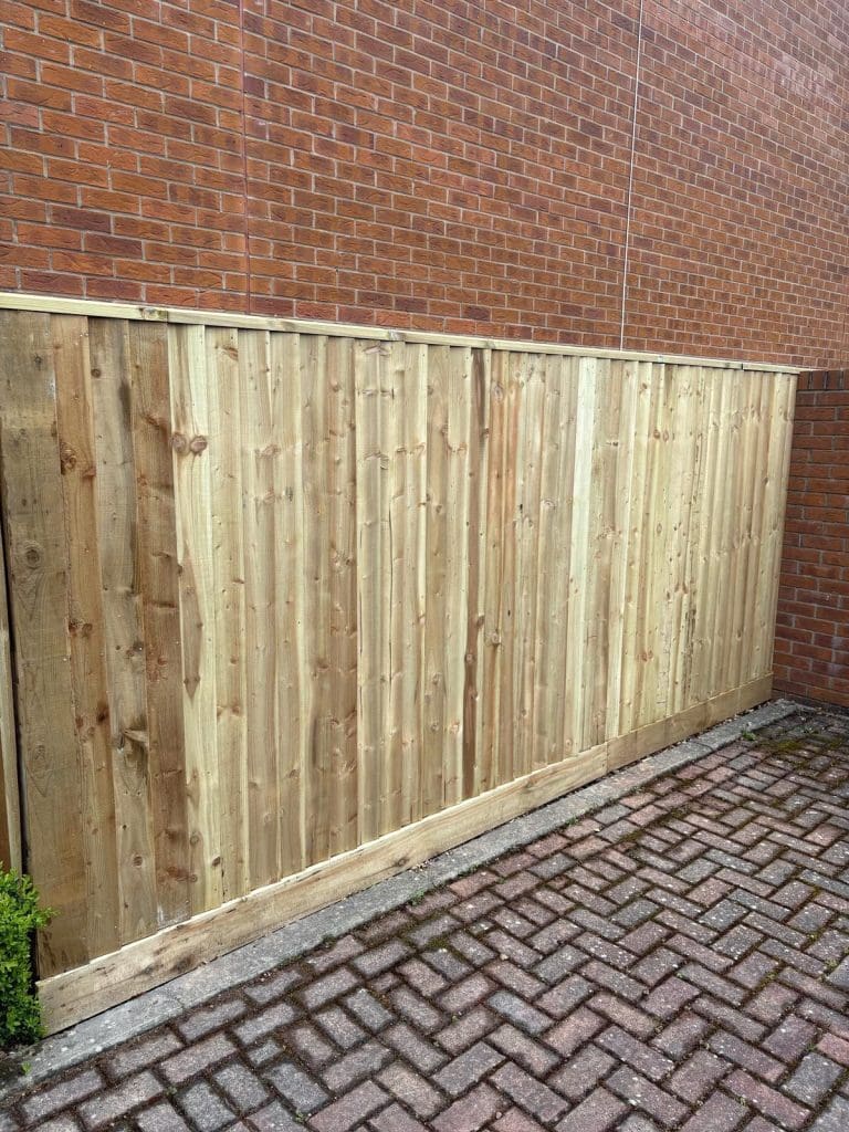 A new wooden fence stands on a paved surface next to a tall red brick wall. The wood appears fresh and unpainted, contrasting with the aged brick background.