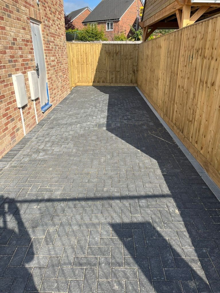 A newly paved driveway with interlocking gray bricks runs alongside a brick house and a tall wooden fence, leading to a closed wooden gate at the far end.