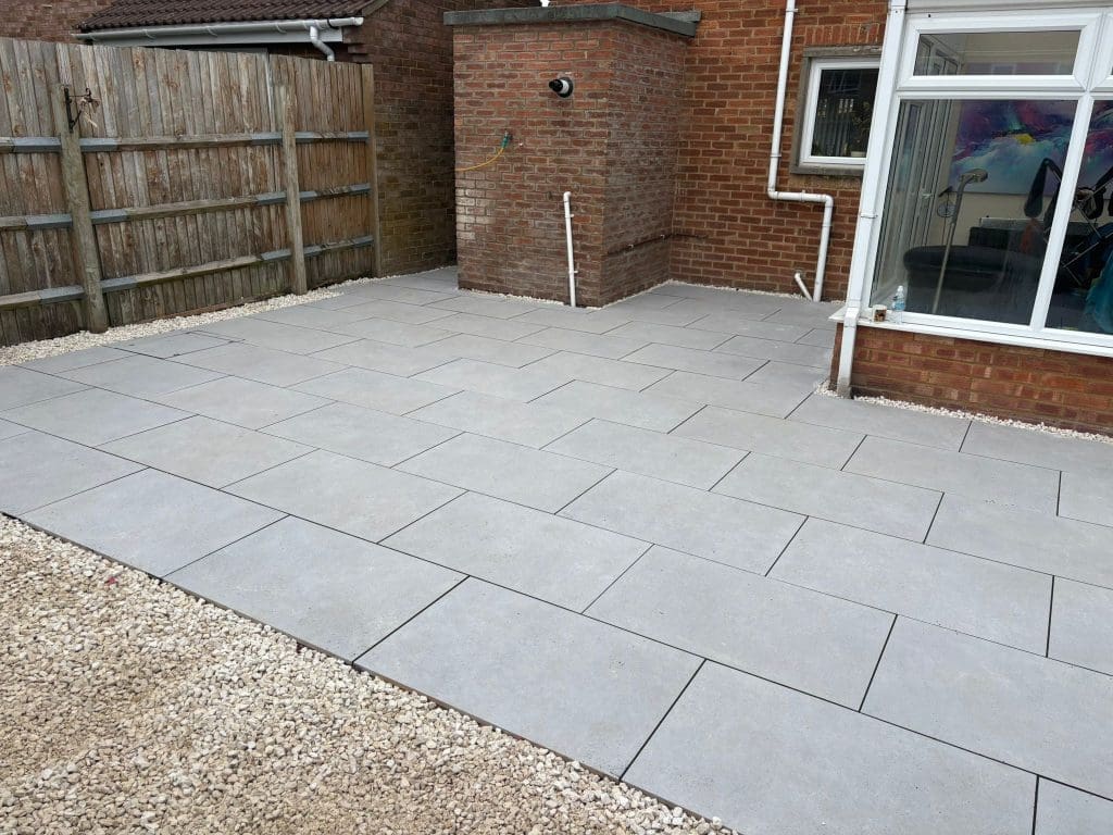 A backyard patio with large, light gray rectangular pavers arranged in a staggered pattern, bordered by light gravel, surrounded by a brick house, wooden fence, and a glass door leading inside.
