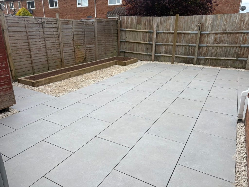 A backyard with large light gray paving slabs, gravel borders, a wooden raised planter along one fence, and wooden panel fencing on three sides. The area is clean and tidy with no plants or furniture.
