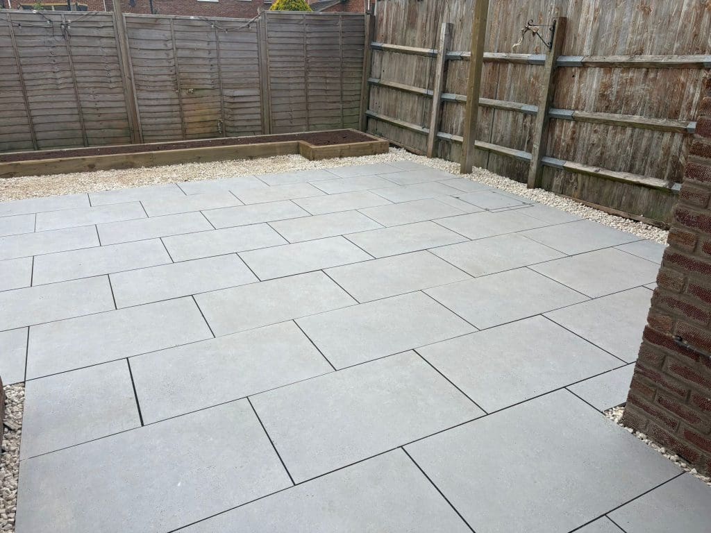 A backyard with large light gray rectangular paving stones laid in a grid pattern, surrounded by gravel and bordered by wooden fences. A small raised garden bed is visible in the background.