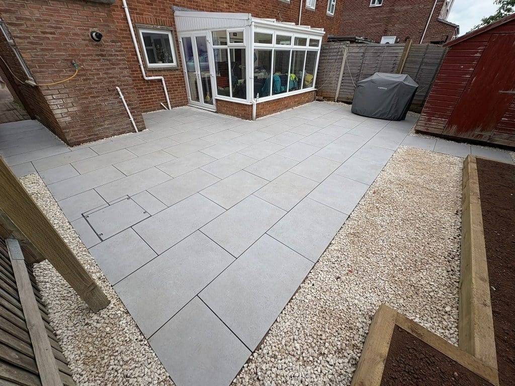 A backyard patio with large light gray paving slabs, surrounded by gravel borders and wooden-raised garden beds. There’s a red shed, a covered barbecue, and a glass conservatory attached to a brick house.