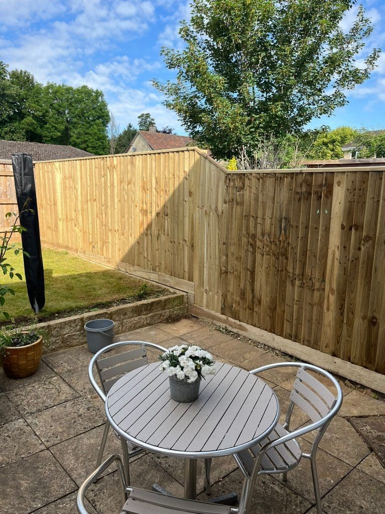 A small patio with a round white table and three metal chairs, featuring a potted white flower centerpiece. The patio overlooks a grassy yard, wooden fence, and leafy trees under a blue sky with clouds.