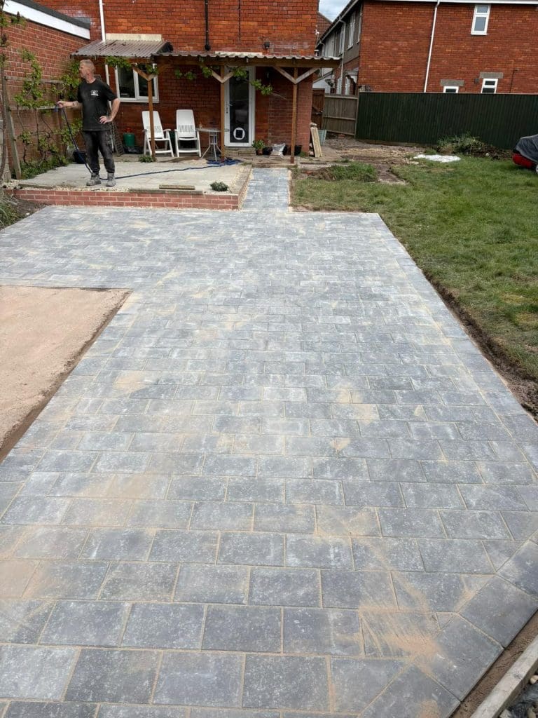 A newly laid grey brick patio extends from a grassy lawn up to a small brick step near a covered patio area, with a man standing to the left side. Some construction materials are visible in the background.