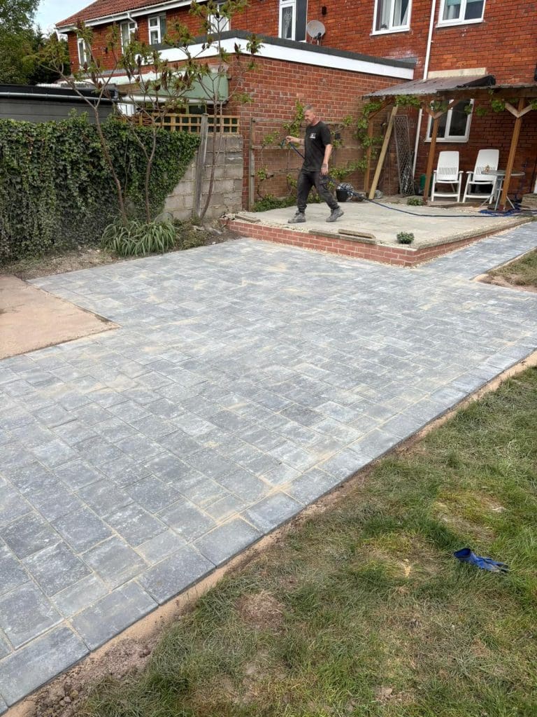 A person works on a backyard patio area with newly laid gray paving stones. There is a grassy lawn nearby and a raised patio with outdoor chairs, next to a brick house and wooden fence.
